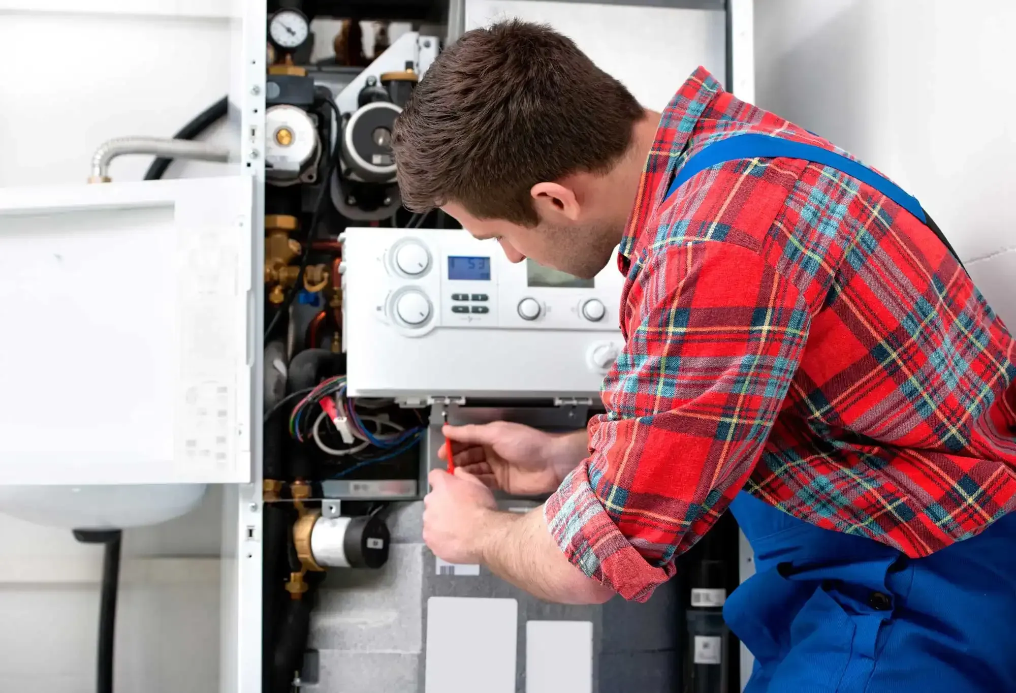 Plumber in red plaid shirt and blue overalls repairing a boiler.