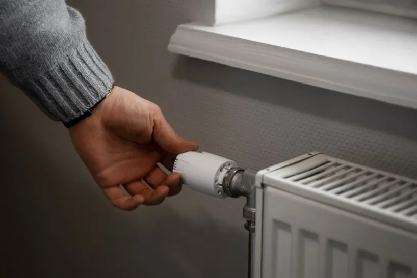 Hand turning a white thermostat valve on a radiator near a window.